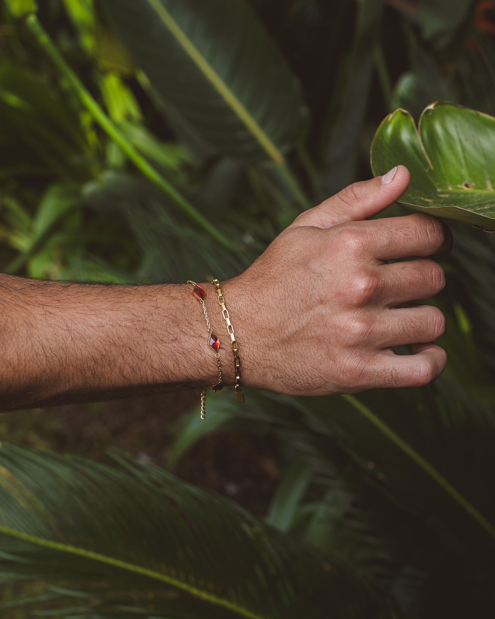 Capri Gold Bracelet with a red gemstone charm worn on a wrist against green leaves
