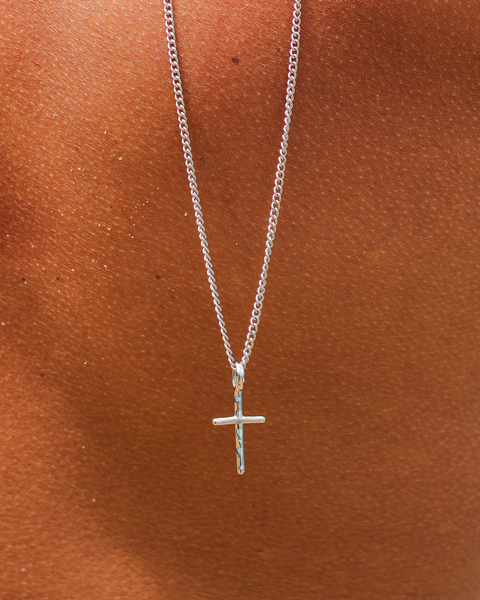 Two silver chain necklaces with a small cross pendant visible at the bottom against a warm brown background.