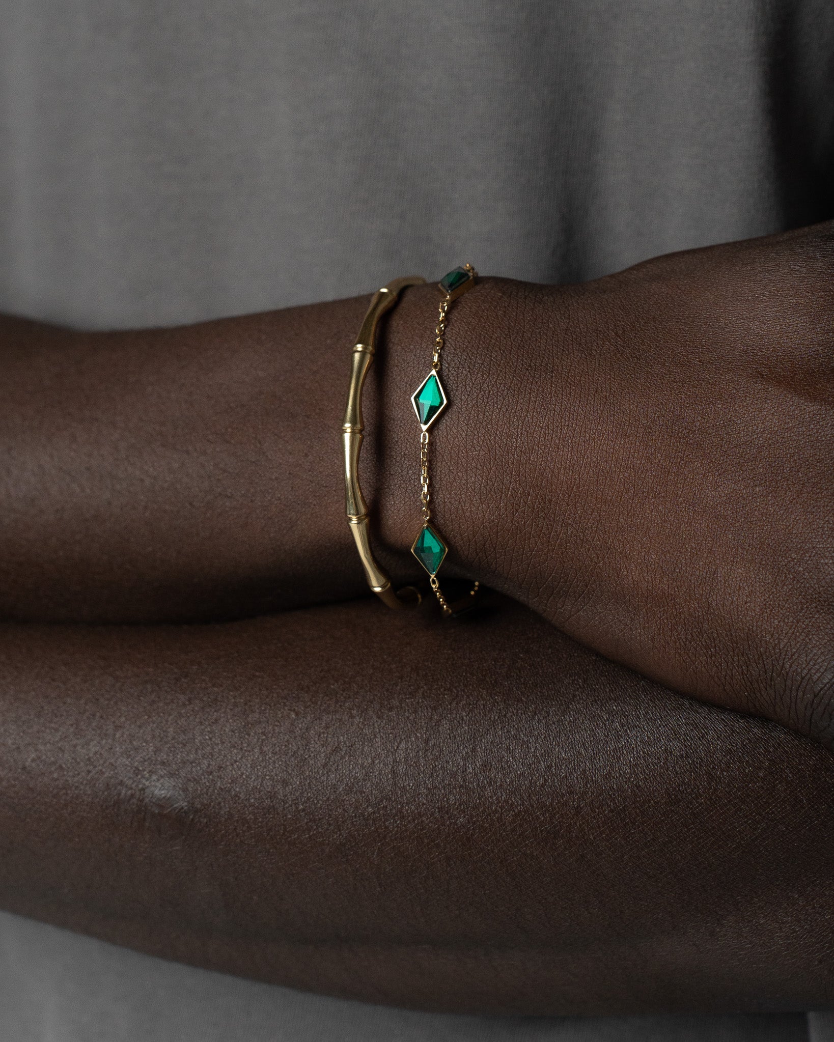 Amberes gold bracelet with diamond shaped emerald stones spaced along a delicate gold chain on dark skin.
