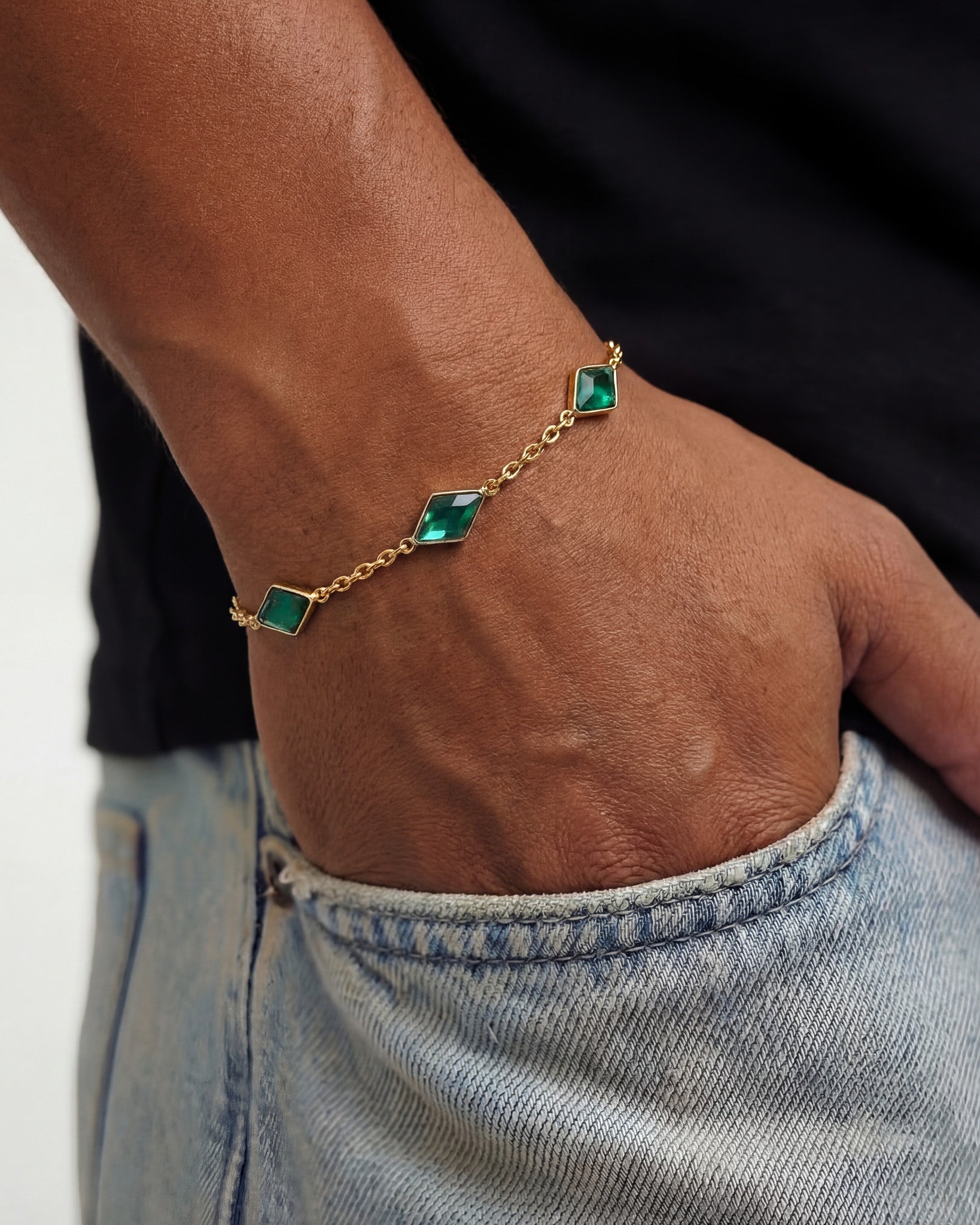 Amberes gold bracelet on a wrist featuring an emerald green square stone and a thin gold chain.