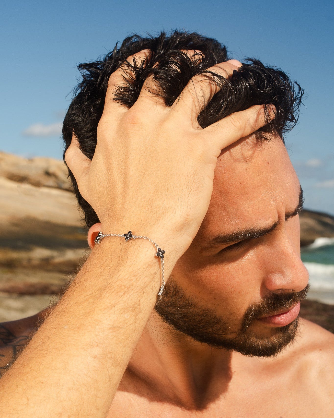 Pulsera plateada Clover con charms de trébol de gema negra en la muñeca de un hombre al aire libre junto a la costa.