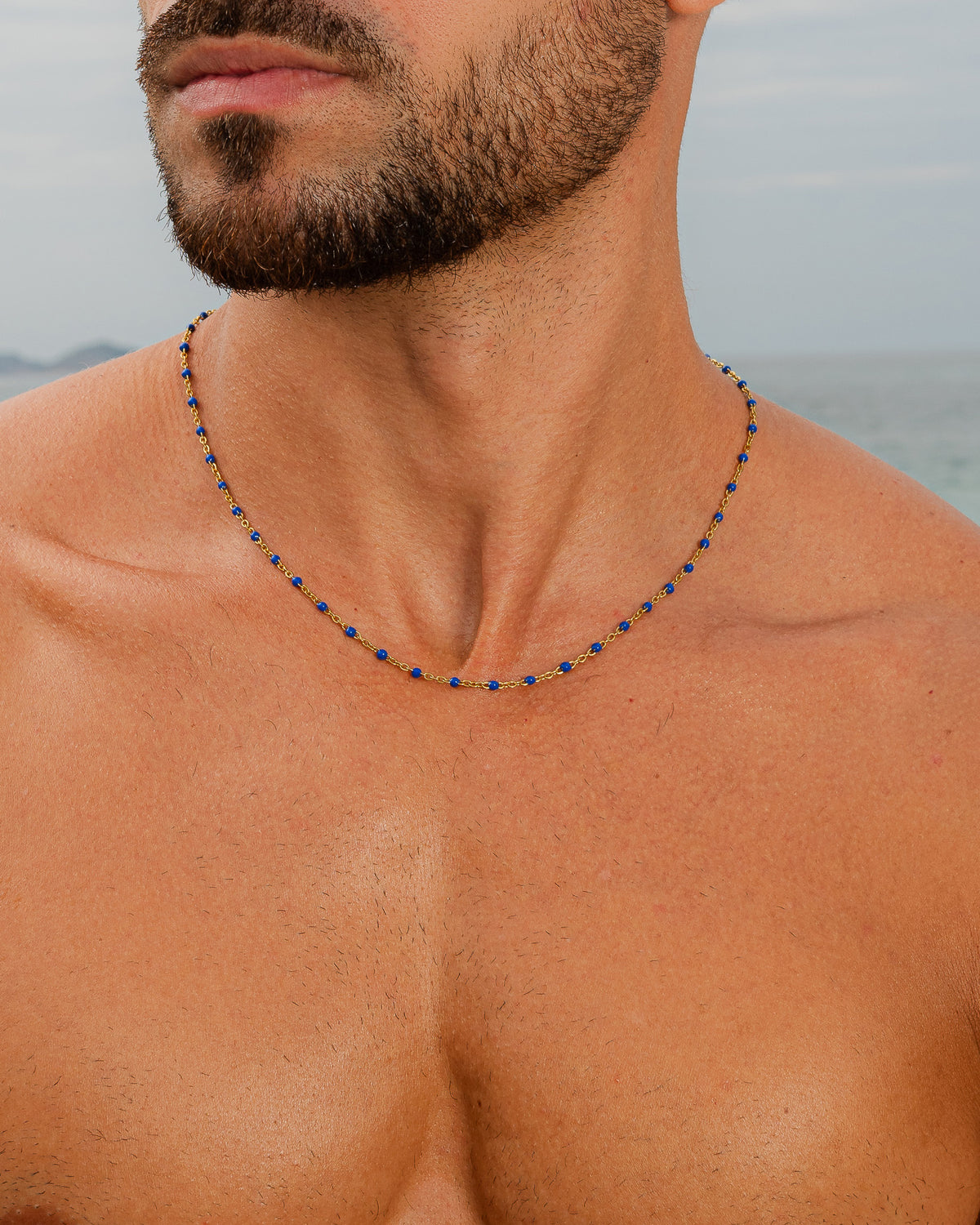 Cadena de oro con cuentas azules en el cuello de un hombre en la playa.