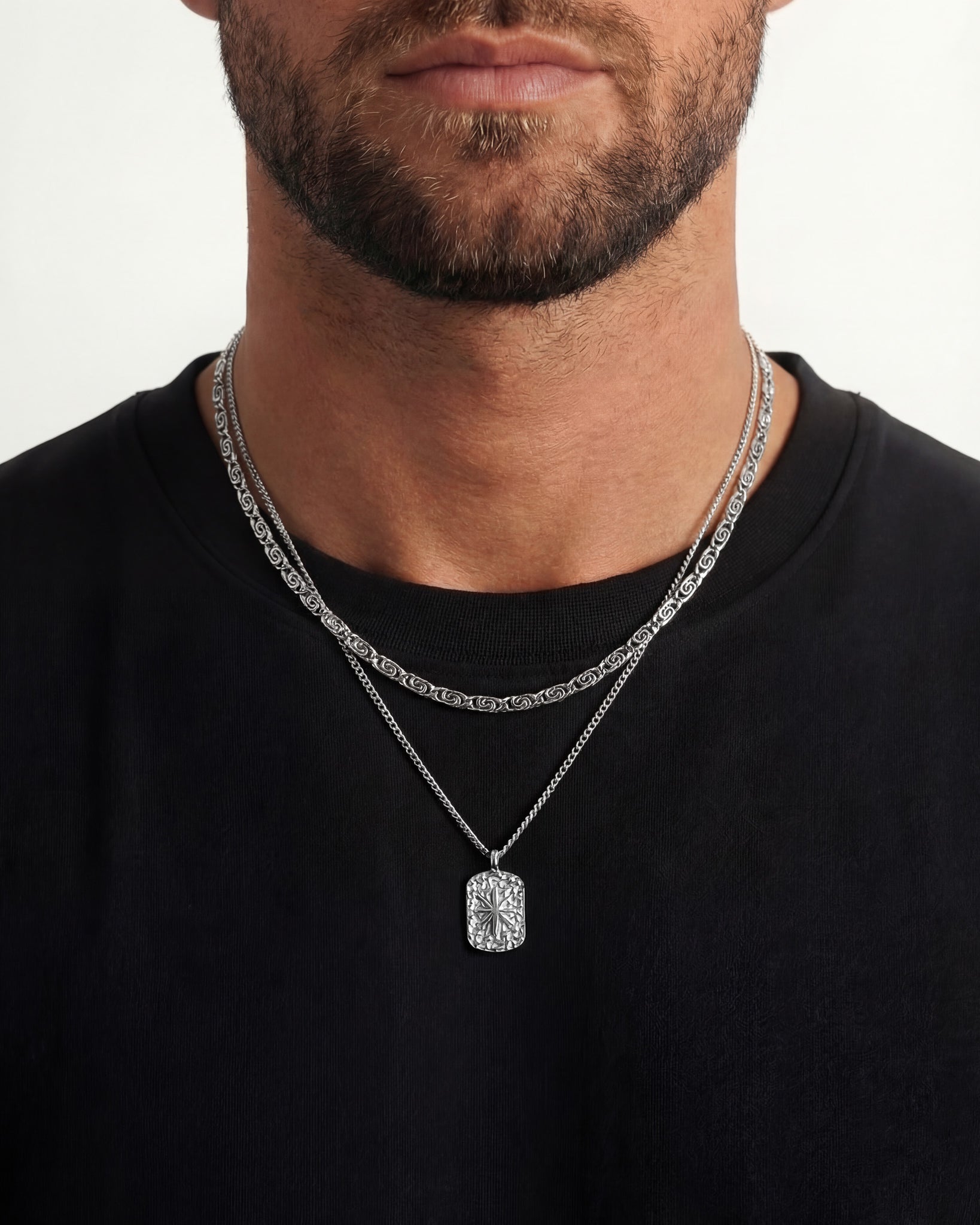 Layered silver necklace with a textured rectangular pendant and a patterned chain on a black shirt