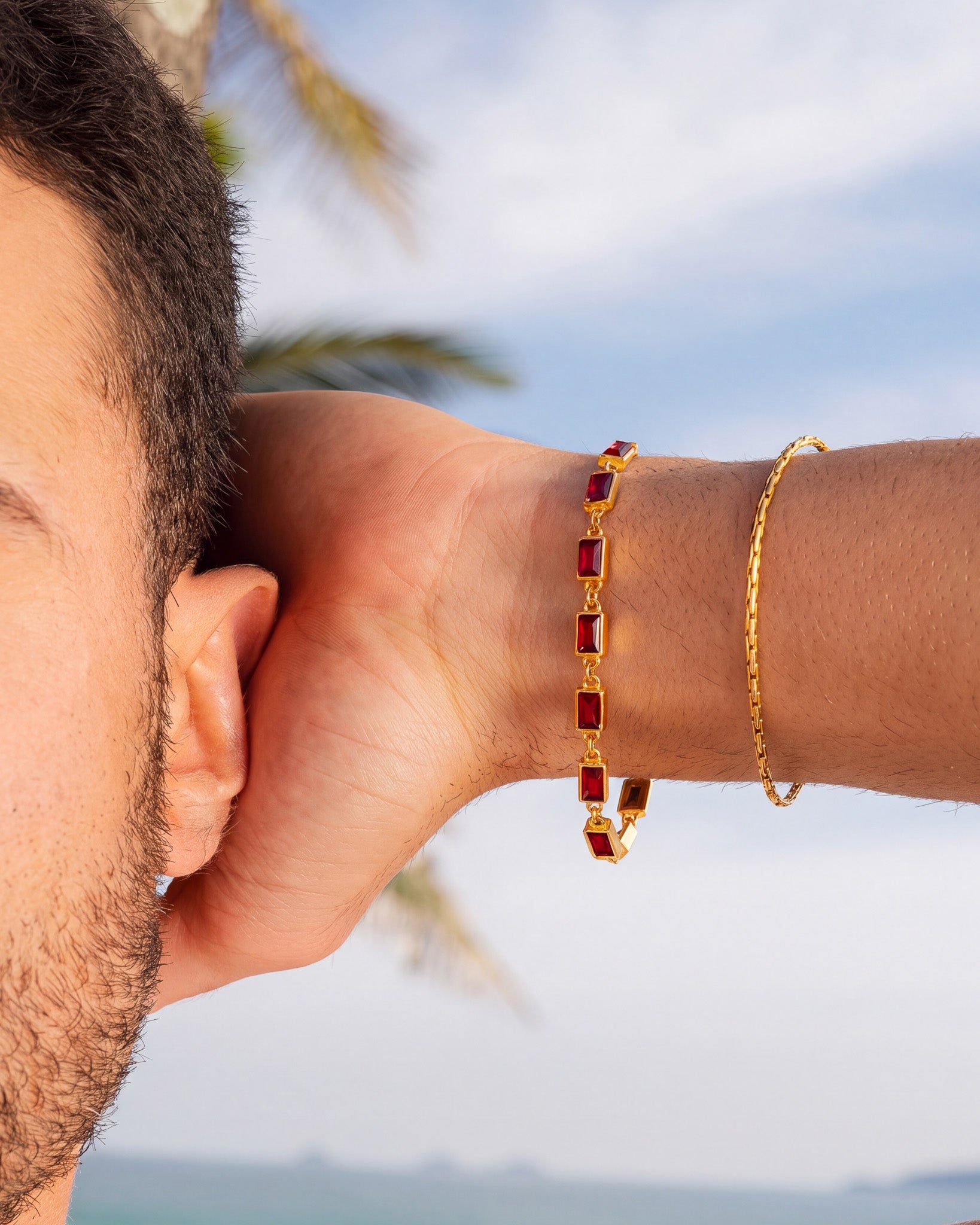 Pulsera OMEN de oro con gemas rectangulares rojas en la muñeca, con el cielo azul de fondo