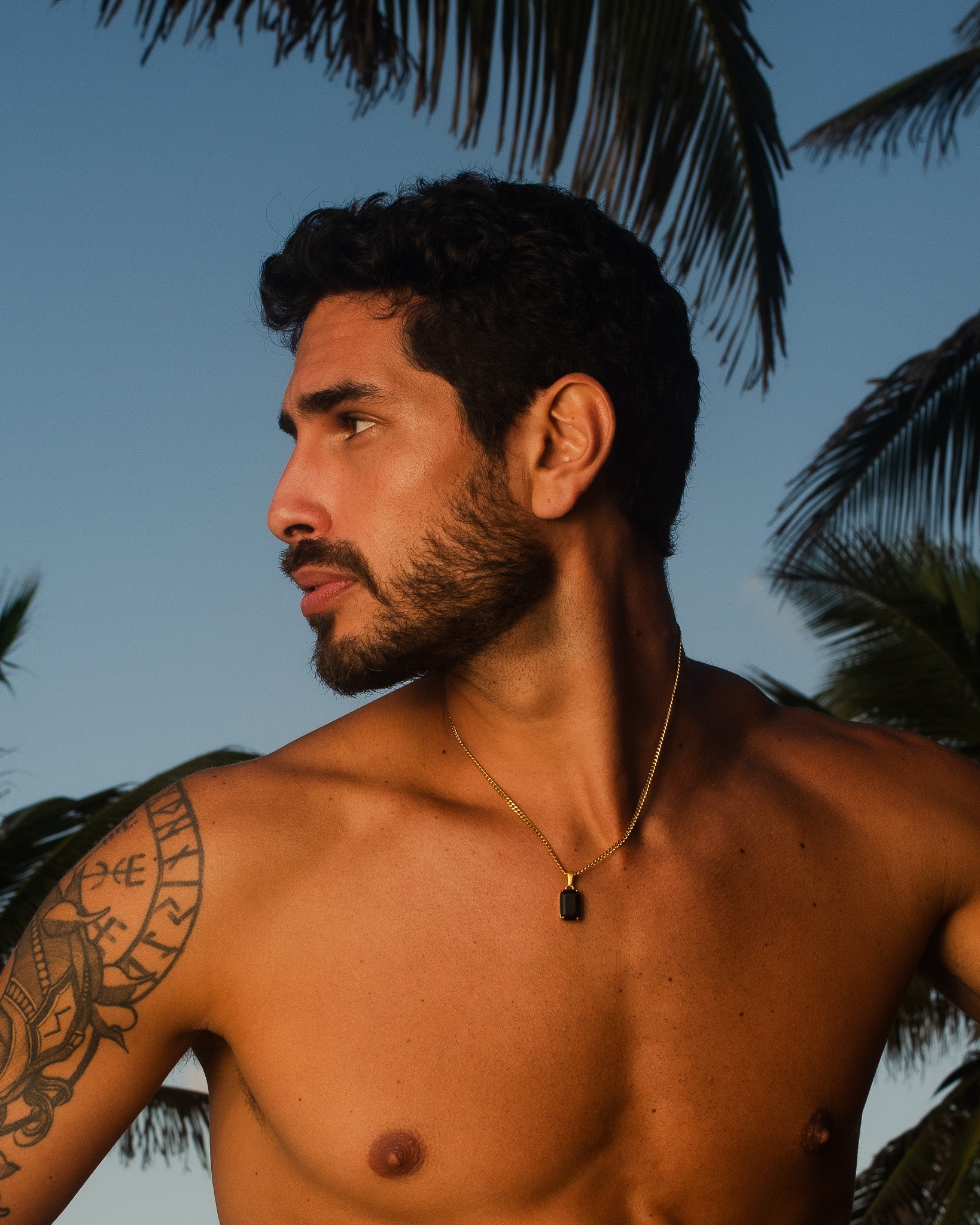 Perfil de un hombre con collar dorado VOLT al aire libre con palmeras de fondo.
