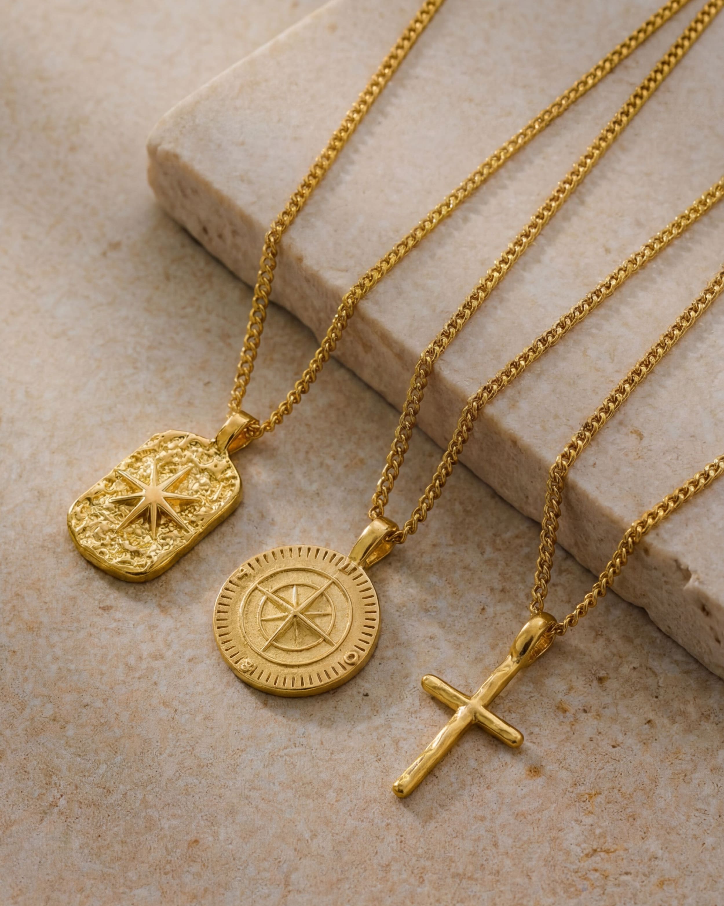 Gold chains spread on a beige surface with rectangular engraved medallion and round compass pendants.