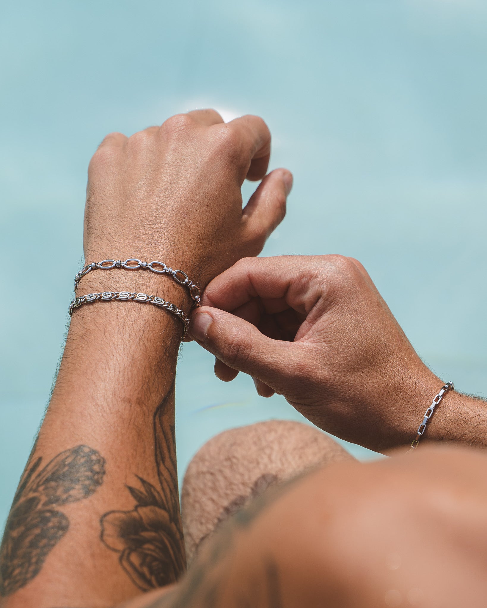 Close-up of a wrist wearing a NUSA Silver Bracelet chain against a pale blue background with visible forearm tattoos.