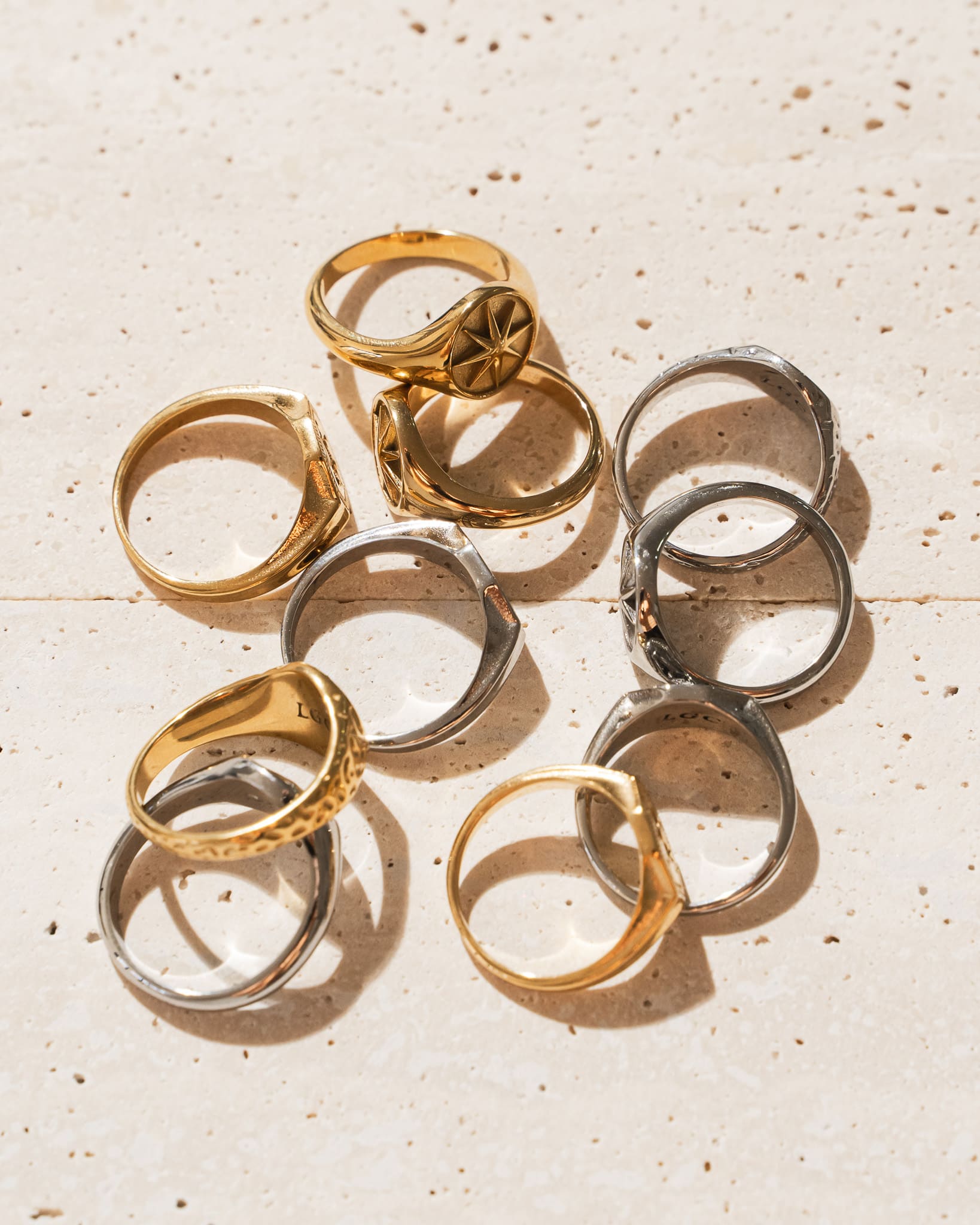 Assortment of Lost Gen Club gold and silver men's signet rings arranged on a travertine stone surface in natural sunlight.