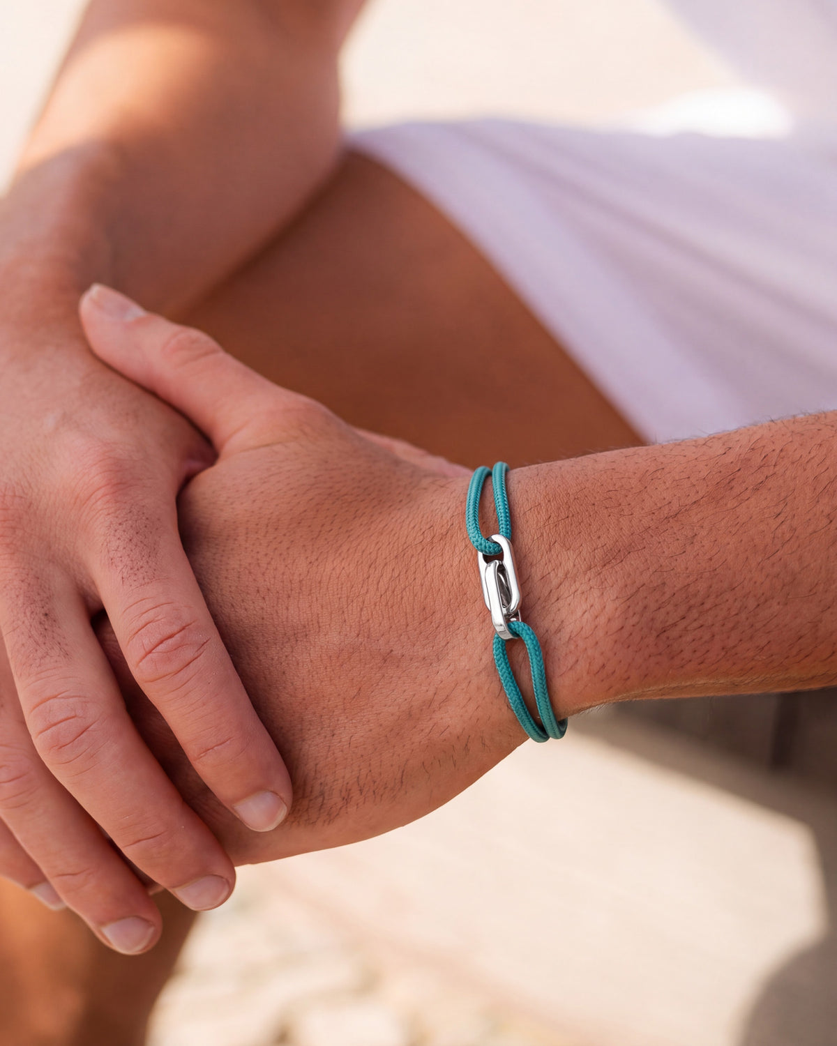 Pulsera plateada Atlantis con cordón azul verdoso en la muñeca, con las manos juntas a la luz del sol