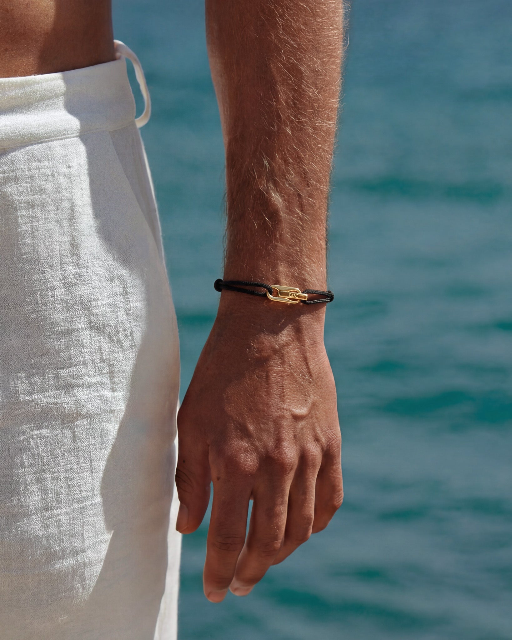 Pulsera dorada Atlantis con cordón negro y cierre dorado en la muñeca junto al mar