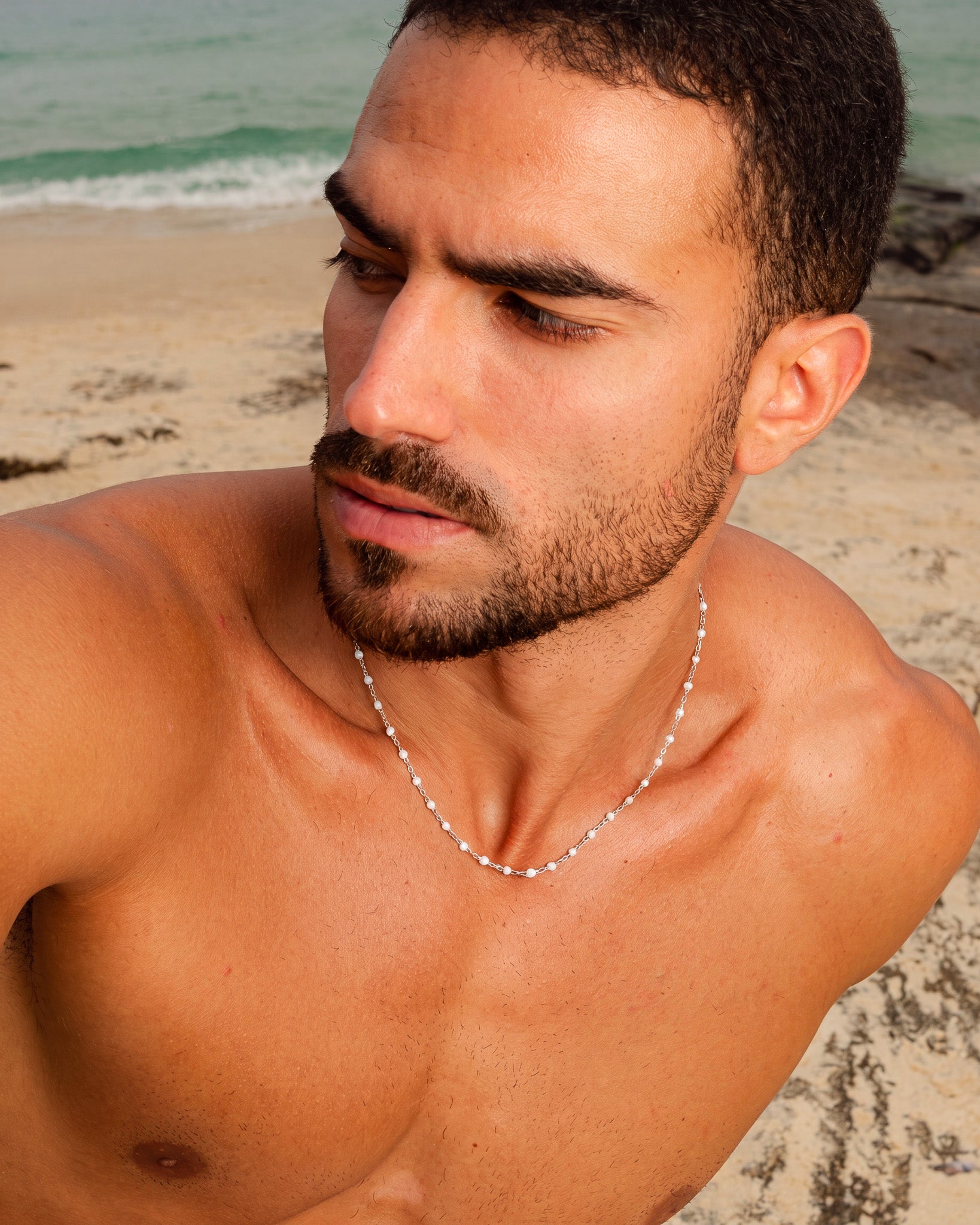 Collar de cadena de plata con cuentas blancas pequeñas sobre el pecho de un hombre en la playa
