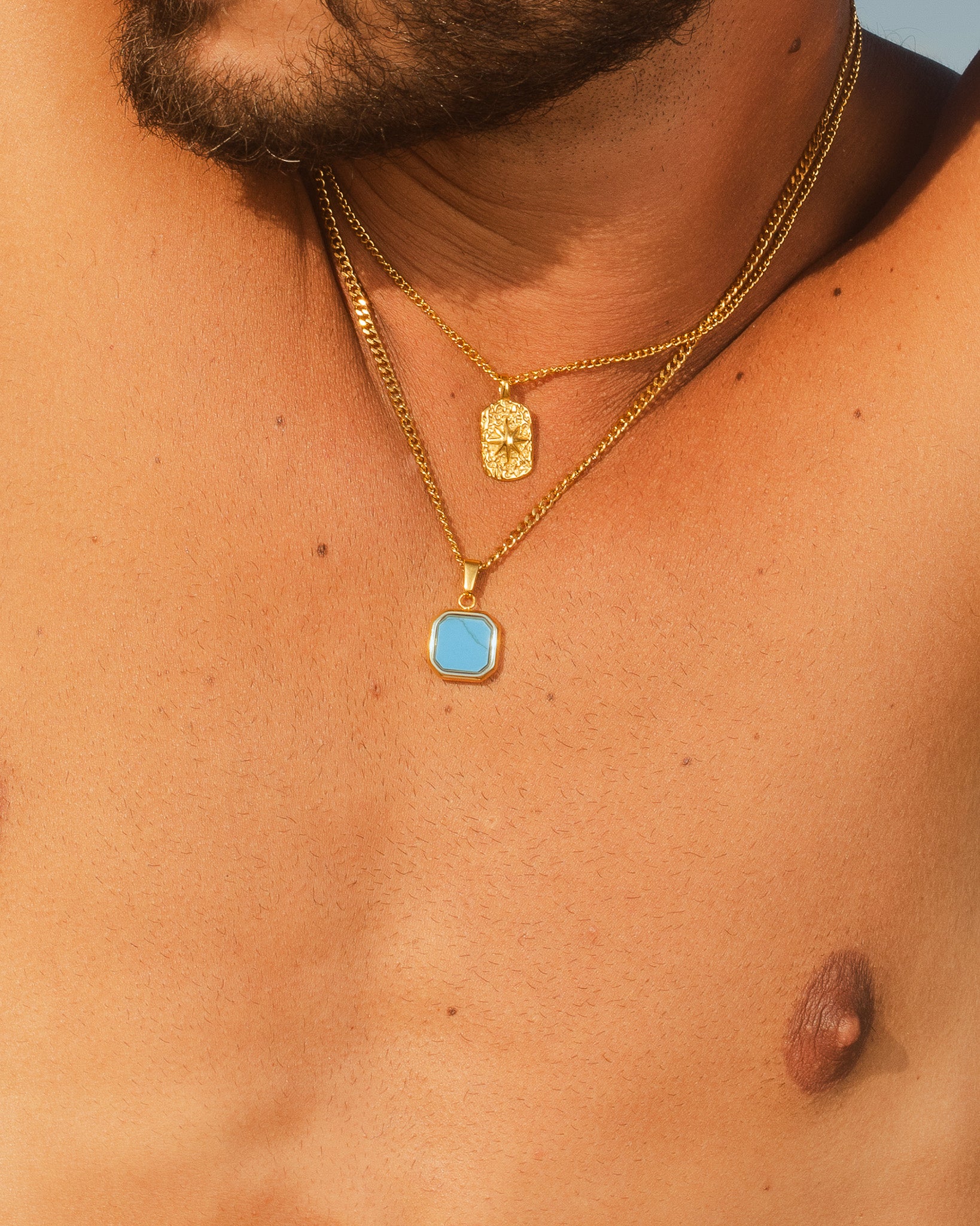 Plano del pecho de un hombre con varios collares dorados Nova con colgante hexagonal turquesa y dos colgantes dorados sobre fondo rocoso