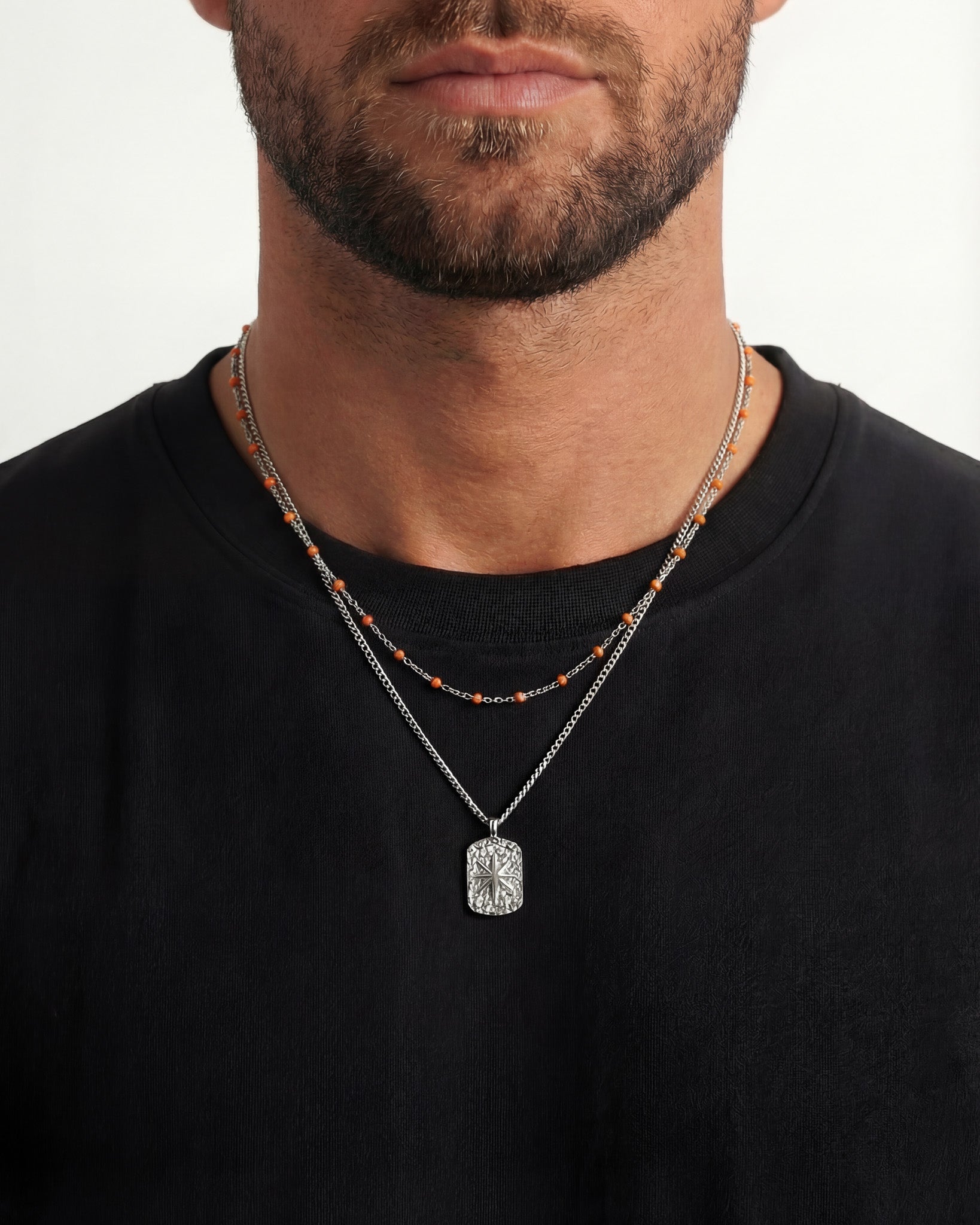 Close up of a man's neck wearing two silver chains; one with orange beads and a textured rectangular pendant.