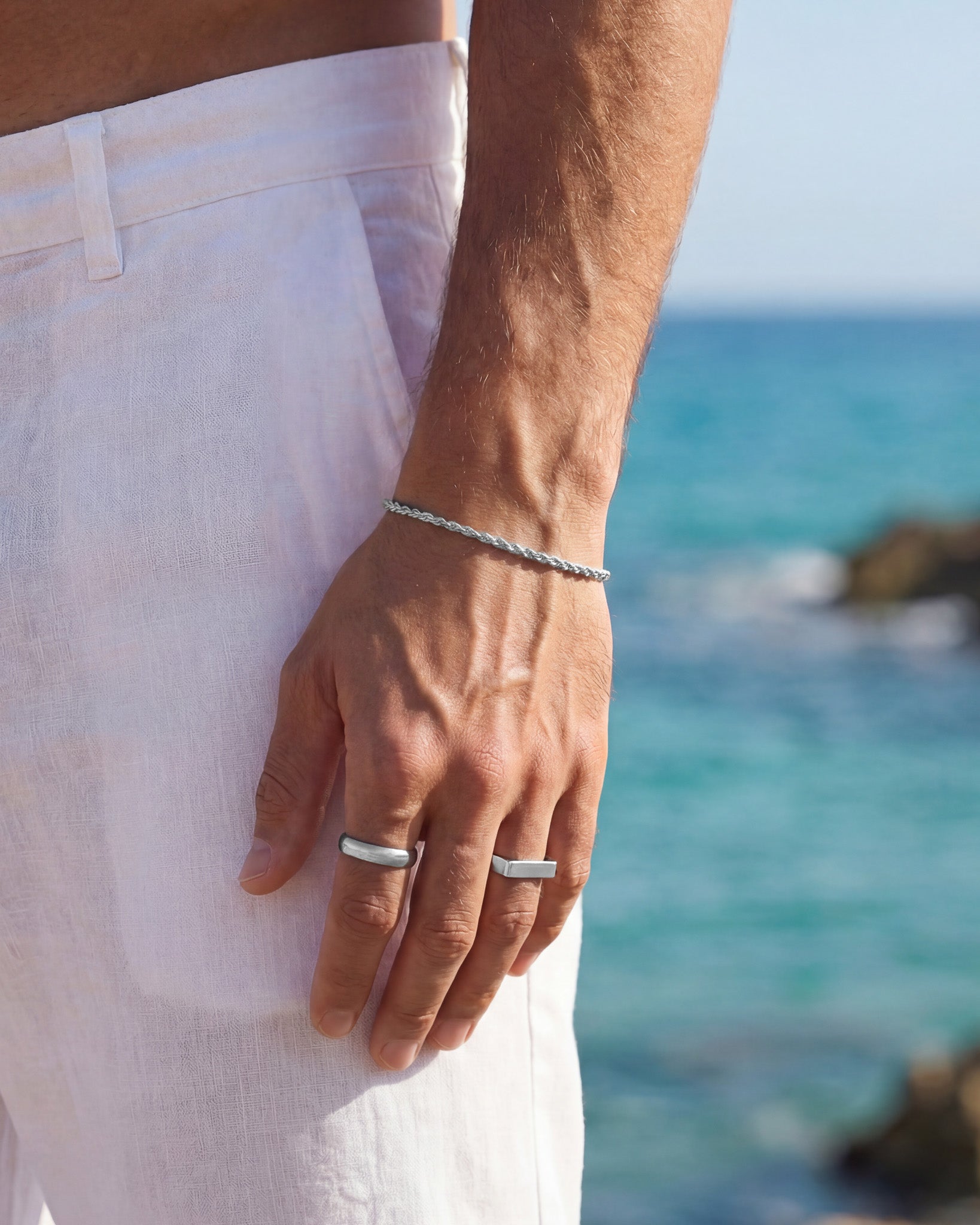 Silver twisted rope bracelet on a man’s wrist with white shorts and ocean backdrop.